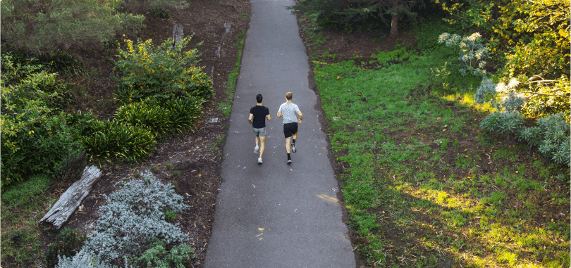 An aerial shot from behind of two people running on an asphalt running trail in a forest